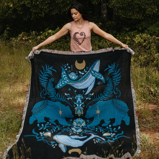 Person holding a large black blanket with blue designs in a forest setting
